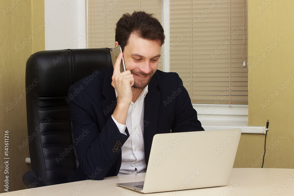 young businessman sitting behind his desk with laptop and talking on mobile phone in office young businessman sitting behind his desk with laptop and talking on mobile phone in office