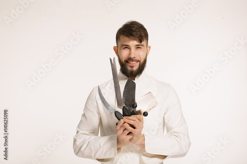 Studio shot of a happy bearded young chef holding sharp knives over white background. Chef with knife. Handsome smiling cheef holding many sharp knives isolated against white background.