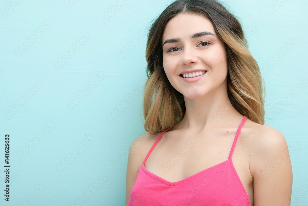 Young girl with long curly hair posing on blue background in studio. She wears shorts, pink T-shirt, pink sunglasses. She keeps hat overhead, holds blue skateboard on booty, looks excited. Young girl with long curly hair posing on blue background in studio. She wears shorts, pink T-shirt, pink sunglasses. She keeps hat overhead, holds blue skateboard on booty, looks excited.