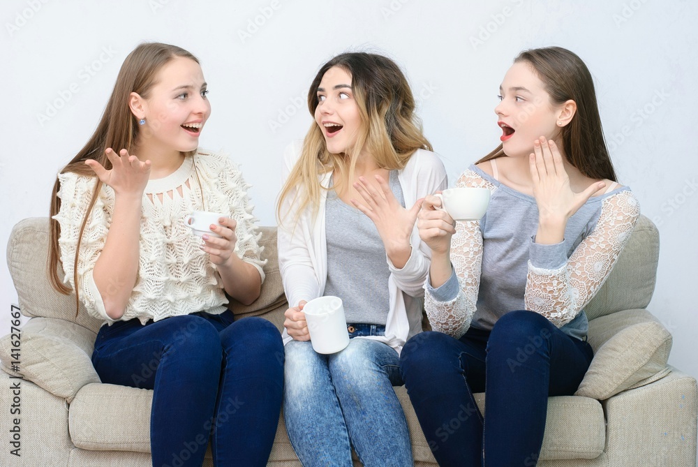 Three happy friends talking and drinking coffee and tea sitting on a couch at home