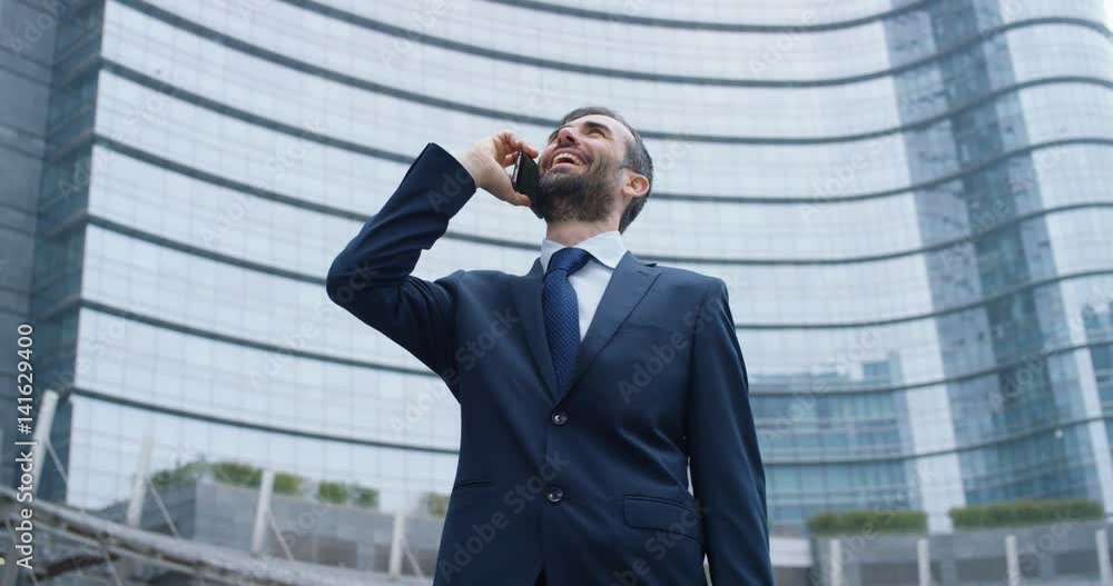 A businessman answering the phone, send messages and smiles for the beautiful job news and in the background you see a skyscraper. Concept: technology, telephony, business trips, business, wall street A businessman answering the phone, send messages and smiles for the beautiful job news and in the background you see a skyscraper. Concept: technology, telephony, business trips, business, wall street