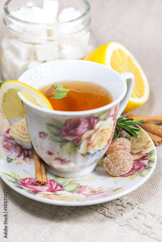 Cup of black or red tea with lemon, rosemary, mint, cinnamon sticks, sugar on grey tablecloth