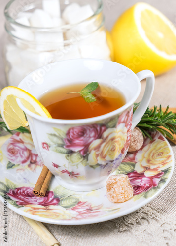 Cup of black or red tea with lemon, rosemary, mint, cinnamon sticks, sugar on grey tablecloth