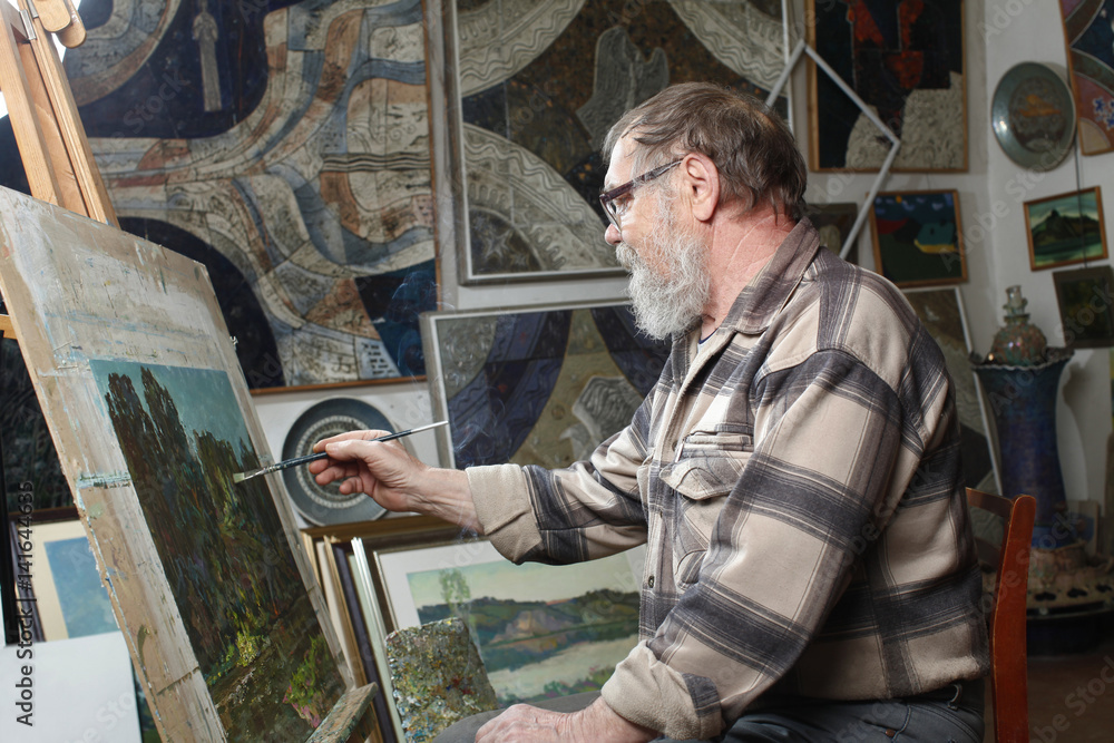 Elderly painter with beard and glasses draws a flowers picture by oil paint in art workshop