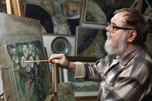 Elderly painter with beard and glasses draws a flowers picture by oil paint in art workshop