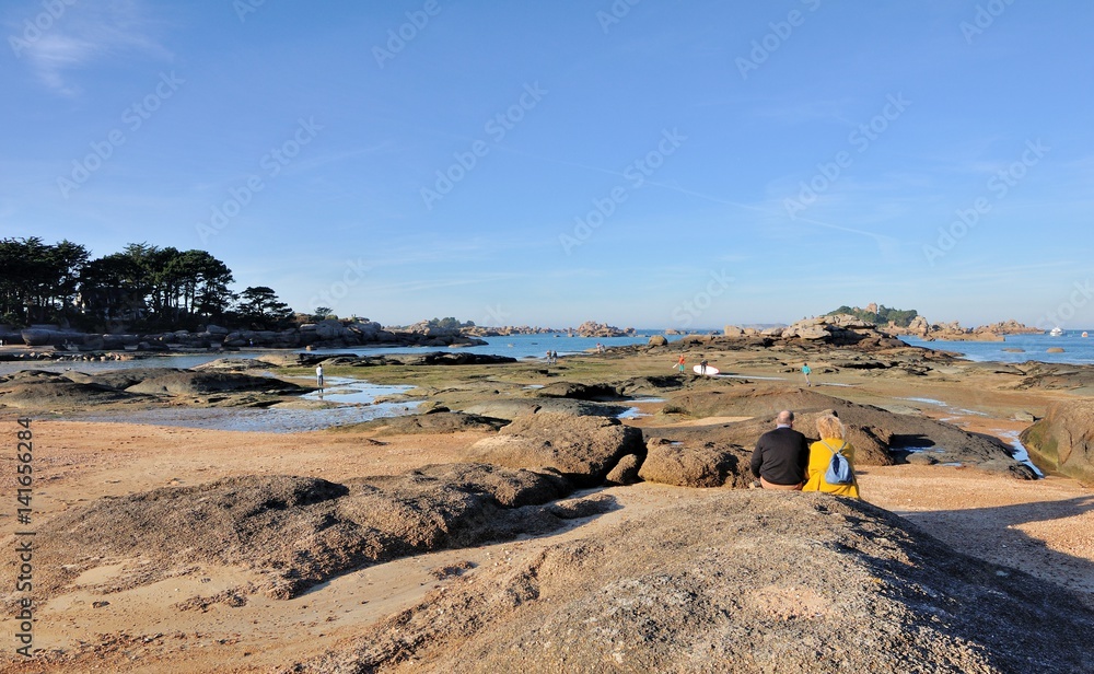 Fototapeta premium Un couple de séniors est assis sur les rochers et admire le paysage de la côte de granit rose en Bretagne