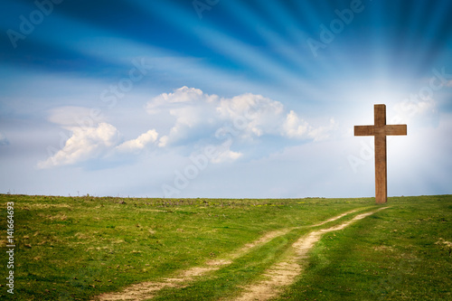 Jesus Christ cross, wooden crucifix on a scene with blue spring sky, green meadows, bright light, rays, clouds. Christian wooden cross on field with green grass and path leading to the cross photo
