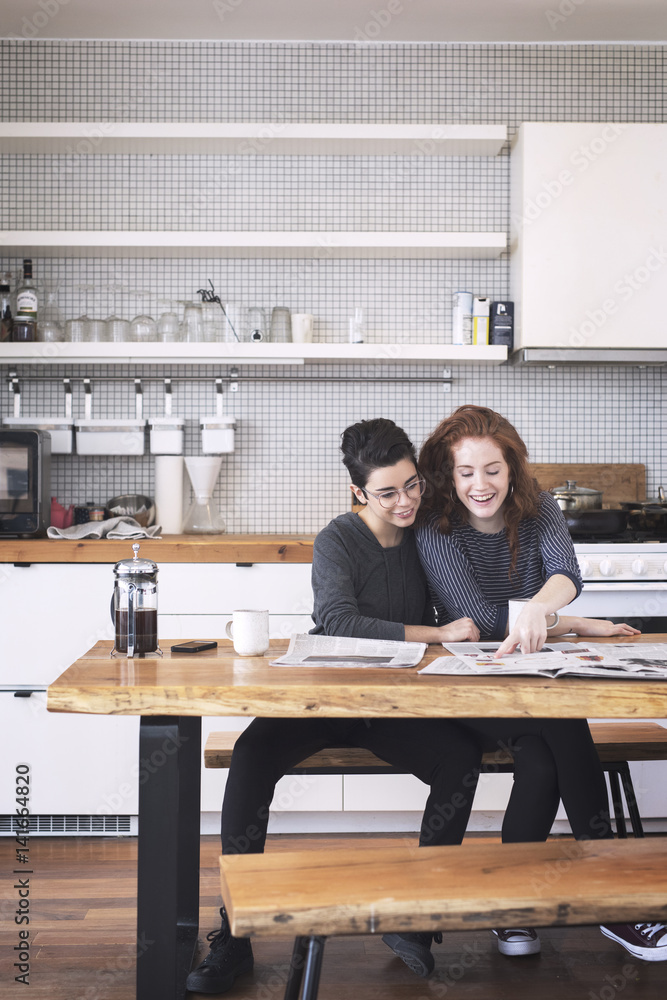 Smiling lesbians couple reading newspaper while sitting at table Smiling lesbians couple reading newspaper while sitting at table