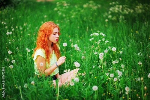 Young beautiful red-haired girl sitting on the green lawn and blowing on a dandelion enjoying summer sun.