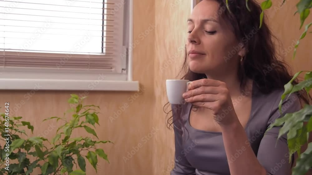 Girl with laptop and coffee. A woman in a gray dress indoors behind a laptop is drinking coffee. Young woman on sofa at a coffee table with laptop. Slow motion.