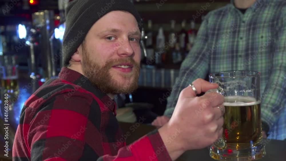 Attractive bearded guy turning back with glass of beer at the pub. Young caucasian man posing with beer mug near the bar counter. Close up of handsome hipster in red checkered shirt and black hat Attractive bearded guy turning back with glass of beer at the pub. Young caucasian man posing with beer mug near the bar counter. Close up of handsome hipster in red checkered shirt and black hat