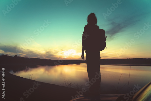 Young woman with backpack standing on car hood at sunset in front of a lake