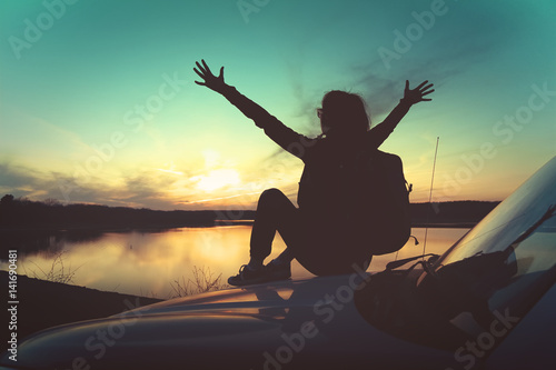 Young woman seating on car hood with hands in the air at sunset in front of a lake