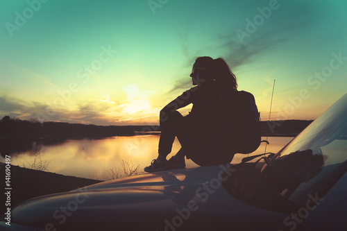Young woman seating on car hood at sunset in front of a lake