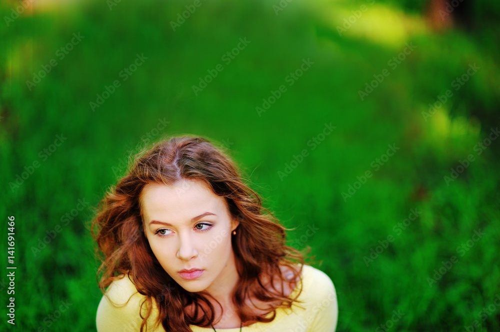 Naklejka premium Beautiful portrait of young pretty pensive brown-haired woman with wonderful hair on a blurry background of bright green grass with the sunlight, close-up.