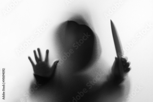 Shadow blur of horror man in jacket with hood knife in his hand.Dangerous man behind the frosted glass.Mystery man.Halloween background.Black and white picture.Blur picture.