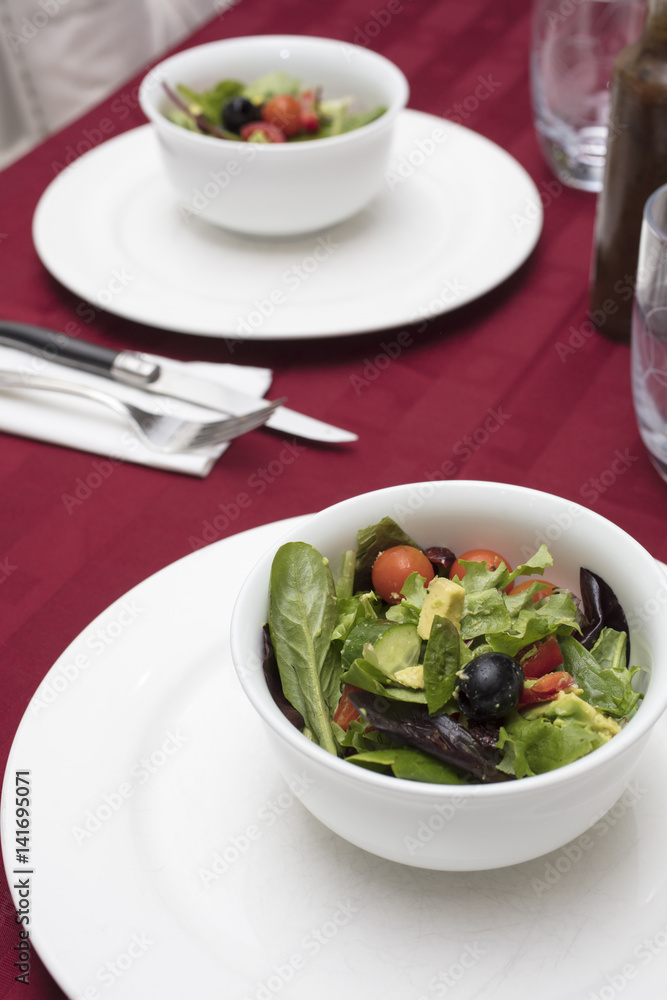 Green Salad with Lettuce, Tomato, Avocado and Olives served in White Dinnerware and Red Table Cloth Green Salad with Lettuce, Tomato, Avocado and Olives served in White Dinnerware and Red Table Cloth