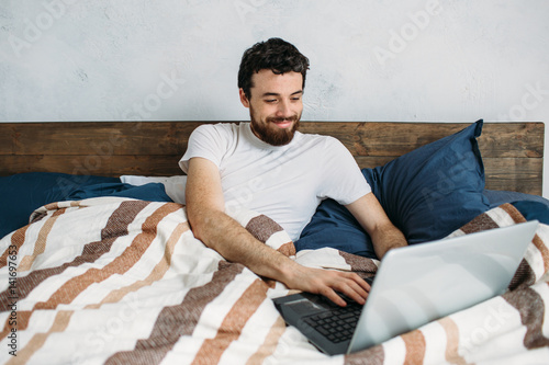 Bearded man lying in morning bed with laptop photo