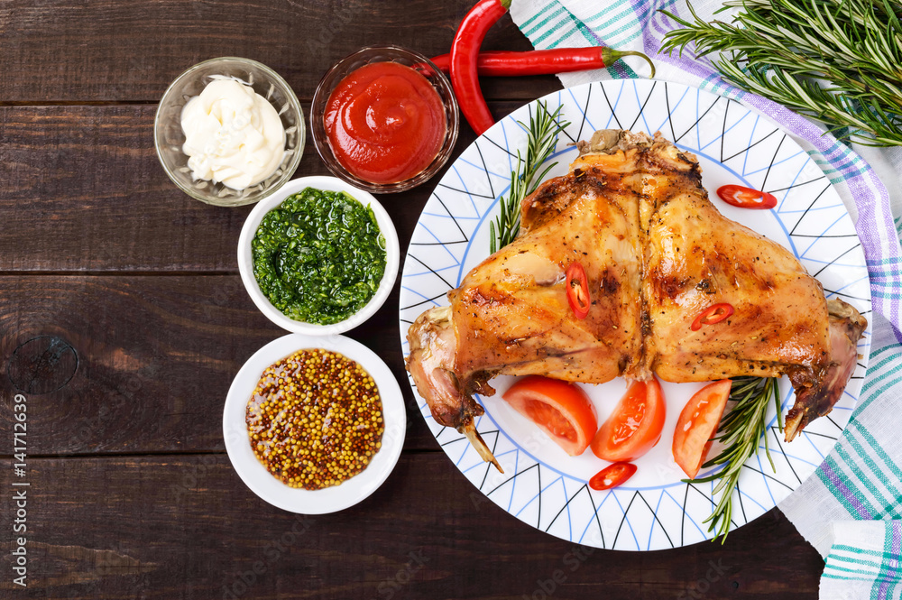 Baked rabbit legs on a plate and different sauces for meat on a dark wooden background. Top view. Baked rabbit legs on a plate and different sauces for meat on a dark wooden background. Top view.