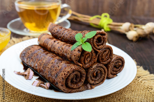 Thin delicate chocolate pancakes, rolled, laid out in a pile on a white plate and a cup of herbal tea