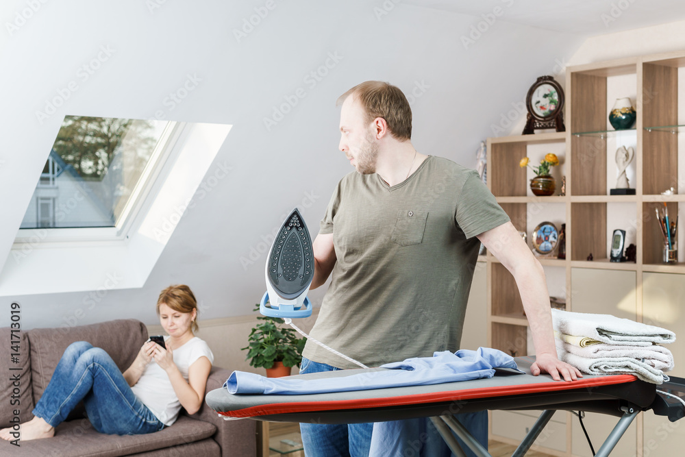 Obraz premium Young woman sitting at the sofa looking at smartphone while her husband is ironing shirt at home.