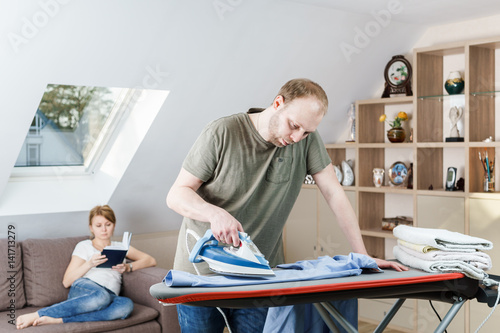 Young woman sitting at the sofa reading the book while her husband is ironing shirt at home.