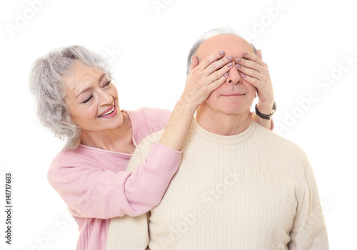 Happy senior woman covering her husband's eyes on white background