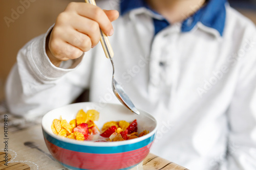 little blond school kid boy eating cereals with milk and berries, fresh strawberry for breakfast