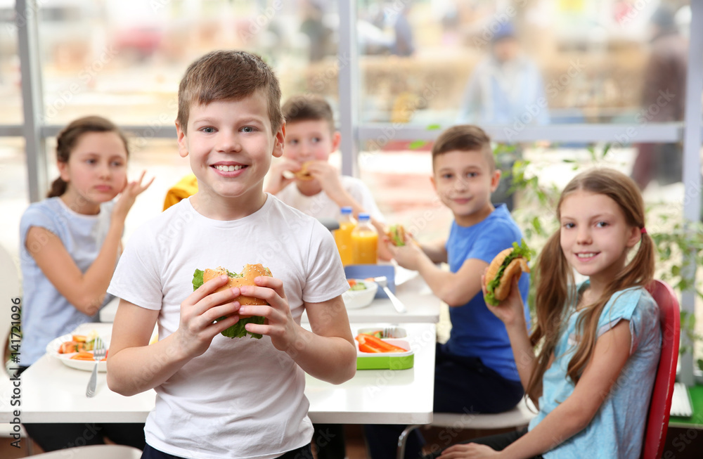 Cute boy eating hamburger and children sitting at table in school cafeteria Cute boy eating hamburger and children sitting at table in school cafeteria