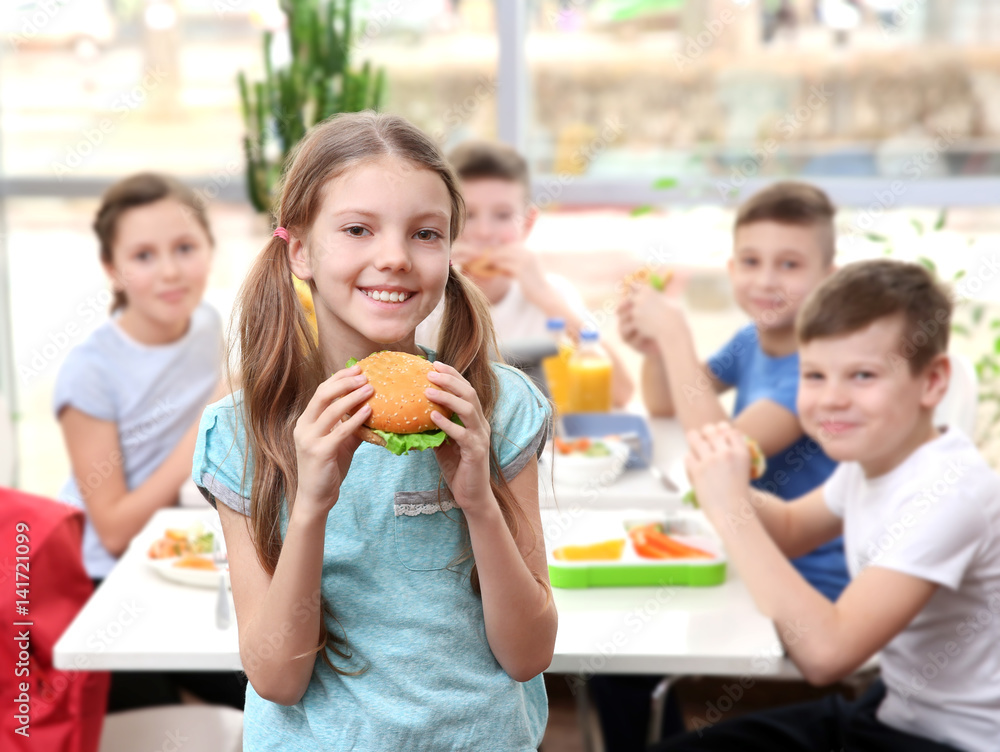 Cute girl eating hamburger and children sitting at table in school cafeteria Cute girl eating hamburger and children sitting at table in school cafeteria