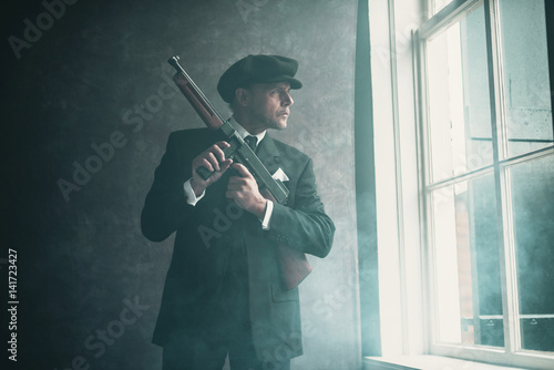 Retro 1920s english gangster wearing flat cap and suit. Standing with gun looking out window.
