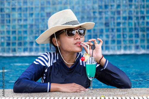 Woman in hat and full body swimsuit relaxing at the pool with cocktail. Woman drinking drink with straw from the glass in the pool. photo