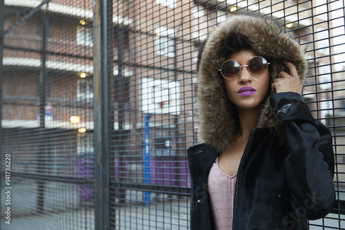 Young woman in fur-lined hood standing by fence, horizontal