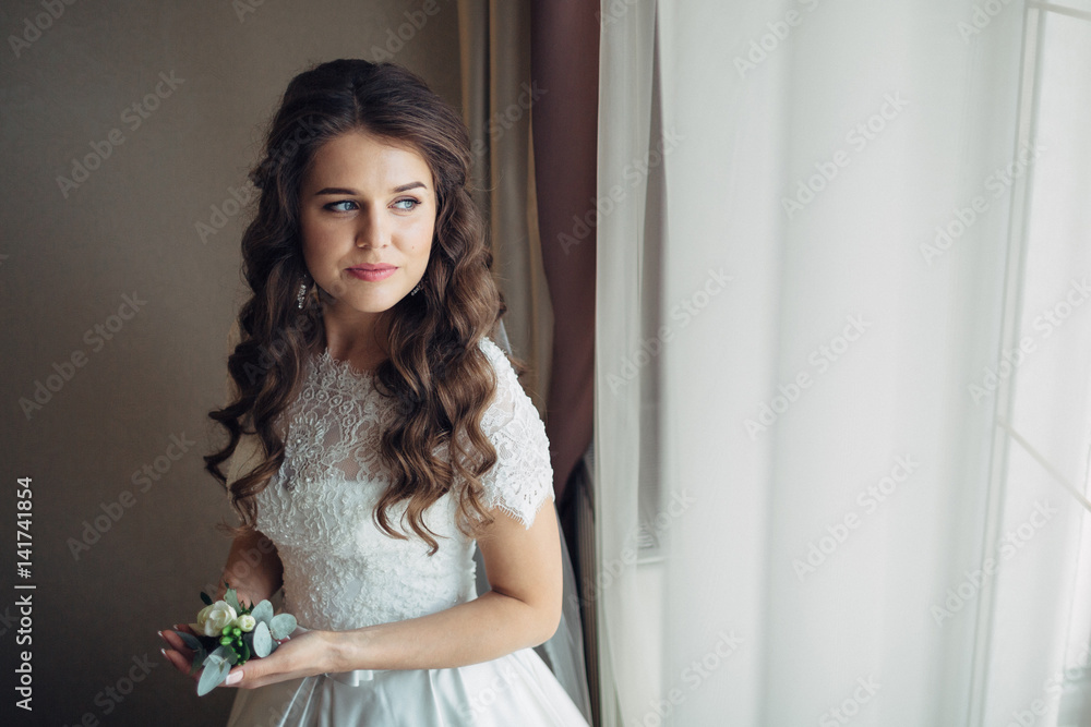 Bride in lace dress stands before bright window with rose boutonniere in her arms Bride in lace dress stands before bright window with rose boutonniere in her arms