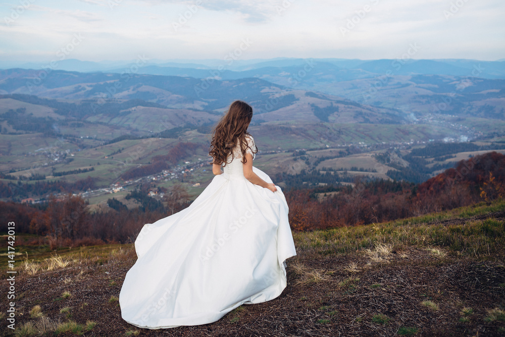 Bride holds her dress in arms walking on high hill with great landcape behind her