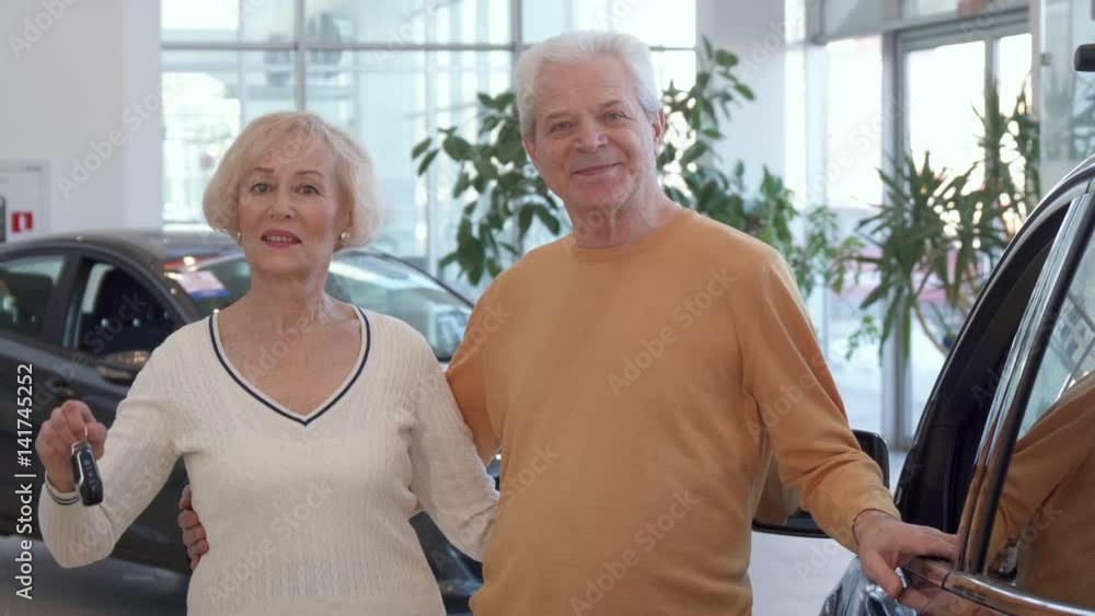 Attractive senior couple buying the car at the dealership. Gray caucasian man embracing his wife against background of new sedan. Aged blond woman showing car key near her husband Attractive senior couple buying the car at the dealership. Gray caucasian man embracing his wife against background of new sedan. Aged blond woman showing car key near her husband