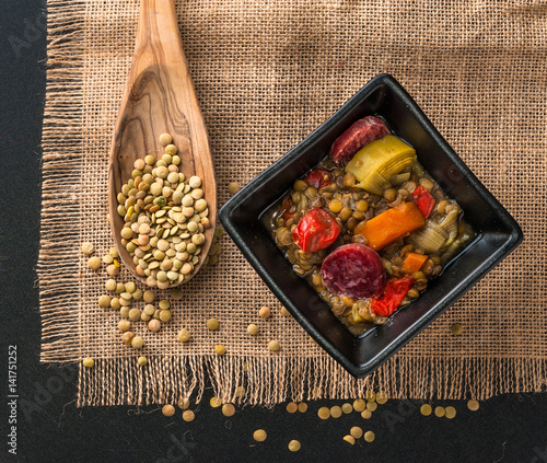colorful lentil soup with carrots, red pepper, leek and chorizo on a black ceramic bowl toether with a wooden spoon full of lentil seeds placed on a rustic table cloth photo