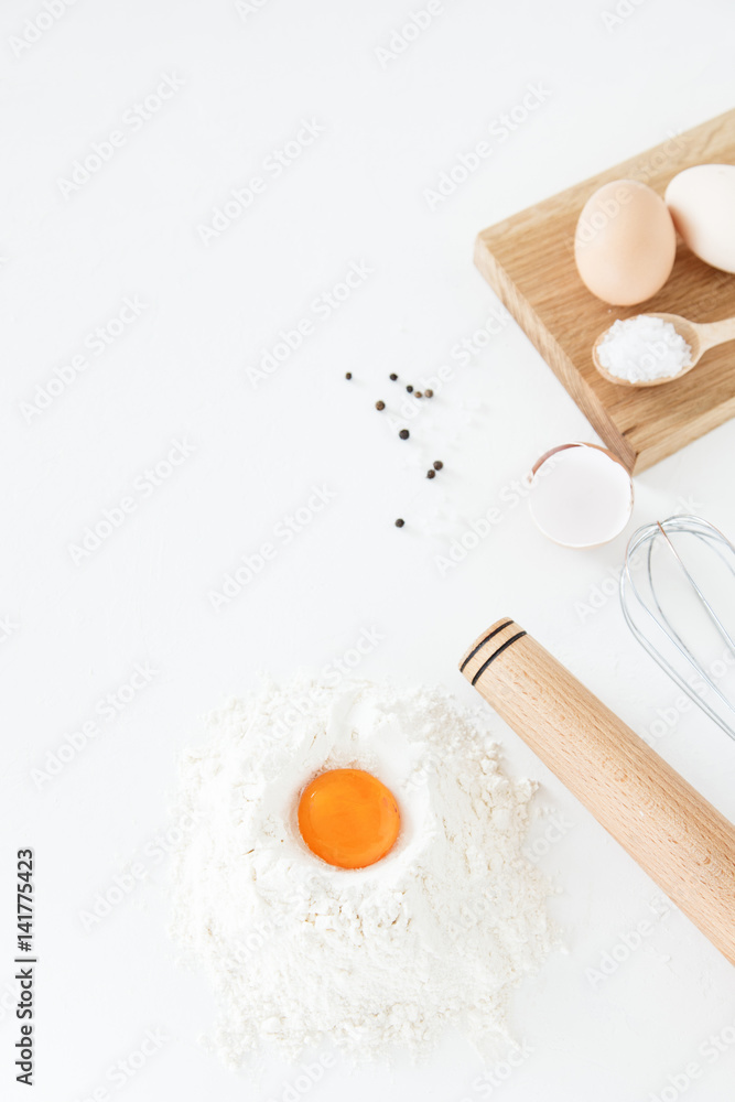 Cooking, culinary accessories and food ingredients lie on a white table. Eggs, flour, salt, pepper, whisk from the mixer, rolling pin and egg yolk. Space for text, daylight. Cooking, culinary accessories and food ingredients lie on a white table. Eggs, flour, salt, pepper, whisk from the mixer, rolling pin and egg yolk. Space for text, daylight.