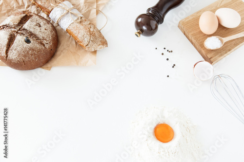 Cooking of homemade bread, black round bread and baguette lie on parchment paper on a white background with flour, sliced bread, paper, salt and eggs. Space for text, daylight.