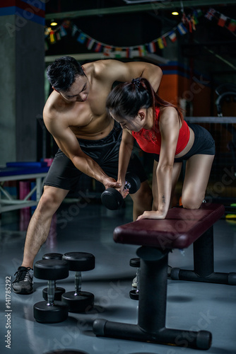 Personal Chinese trainer working with his client in gym. Male Chinese personal trainer teaching woman to make right fitness move with the dumbbell.