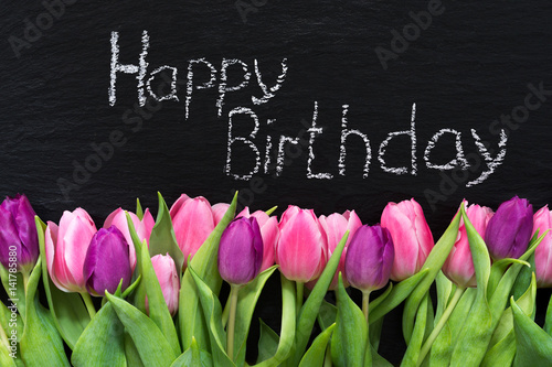 Happy Birthday mit weißer Kreide auf einer Schiefertafel geschrieben und mit Blumen ausgelegt