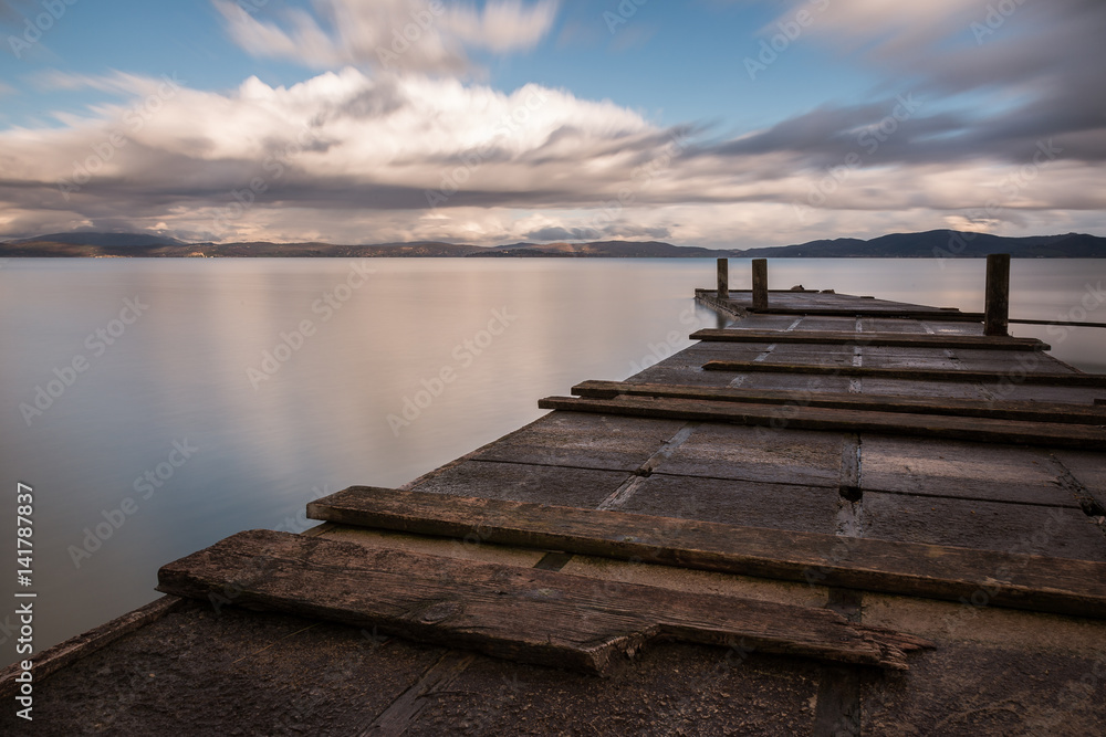 A very close, low view of a curved pier, with still water and a sky with clouds in motion, captured as long exposure