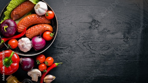 Grilled sausages in a pan with fresh vegetables. Wooden surface. Top view.