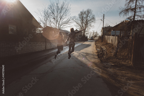 a young girl and a guy in a brown leather jacket running, jumping on the road in the village at sunset on a clear spring day, holding hands