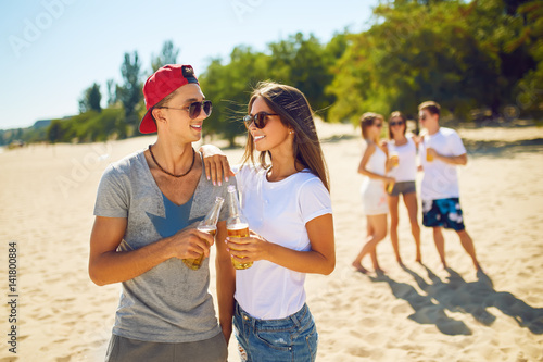 boy and a girl are holding bottles of beer in hands and smiling .Group of friends hanging out with beer at the beach.  Excellent sunny weather. Beautiful figures. Super mood. Summer concept   © maxbelchenko