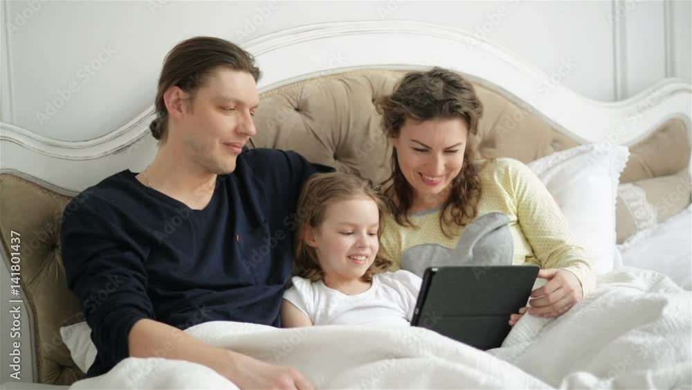 Modern Family is Using Electronic Gadget Lying on the Bed Together. Handsome Man, Brunette Woman and Cute Little Girl is Looking on the Screen of the Tablet Relaxing at Home. Modern Family is Using Electronic Gadget Lying on the Bed Together. Handsome Man, Brunette Woman and Cute Little Girl is Looking on the Screen of the Tablet Relaxing at Home.