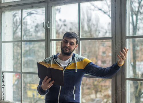 Bearded poet man reads a book with an expression gesture. Big window on background.  photo