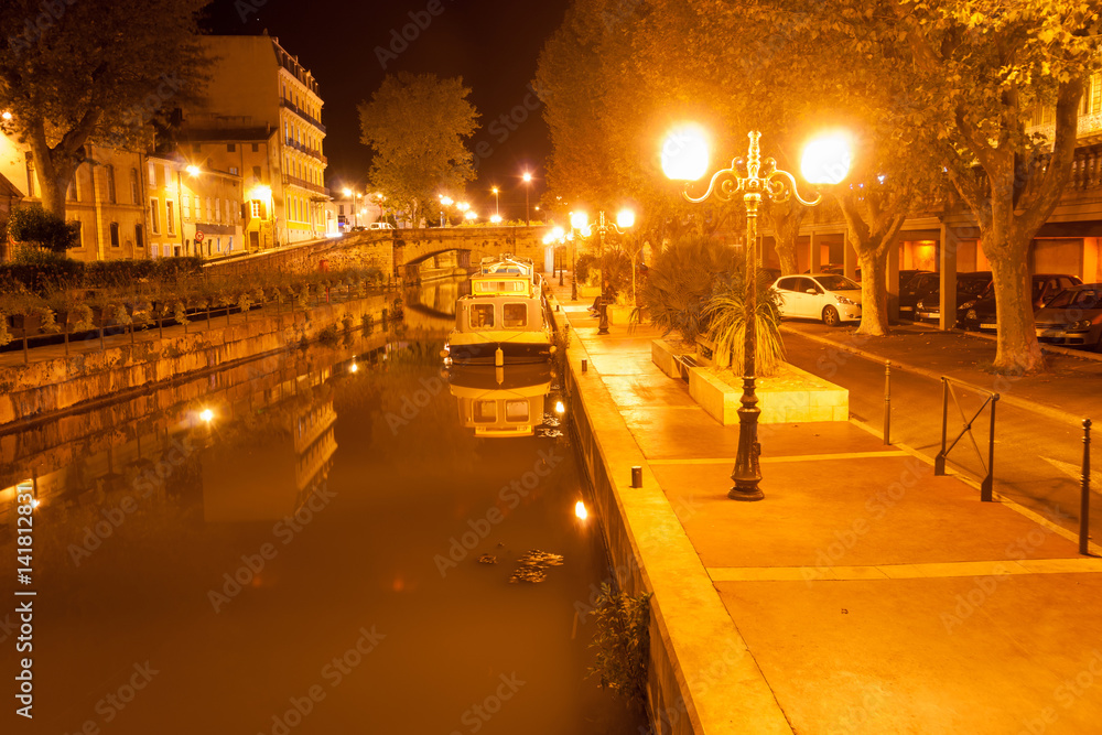 Canal boats moored alongside on Canal de la Robine at night under golden light of street lamps Narbonne, France.