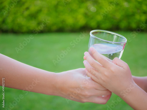 Woman hand giving glass of fresh water to child on the green grass background