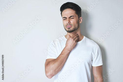 Young man having sore throat and touching his neck, wearing a loose white t-shirt against light grey background. Hard to swallow photo